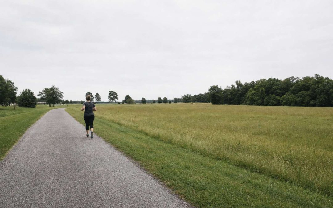 A beginner runner seen from far off, small in the frame, jogging along a wide open greenway